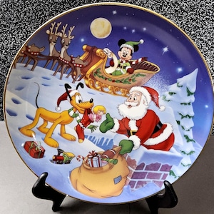 May include: A decorative plate featuring a Christmas scene with Mickey Mouse in a sleigh pulled by reindeer, Pluto, and Santa Claus. The plate has a gold rim and a blue background with a full moon and stars. Santa is holding a sack of gifts.