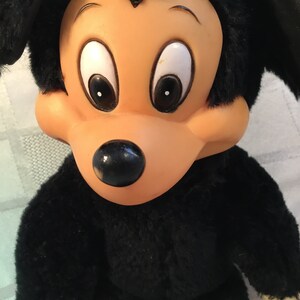 Puede incluir: Un peluche con un personaje de dibujos animados con un cuerpo negro y orejas grandes y redondas. El personaje tiene una cara bronceada con ojos grandes y blancos y una nariz negra. El pelaje del juguete es negro y suave, con una expresi&oacute;n facial detallada.