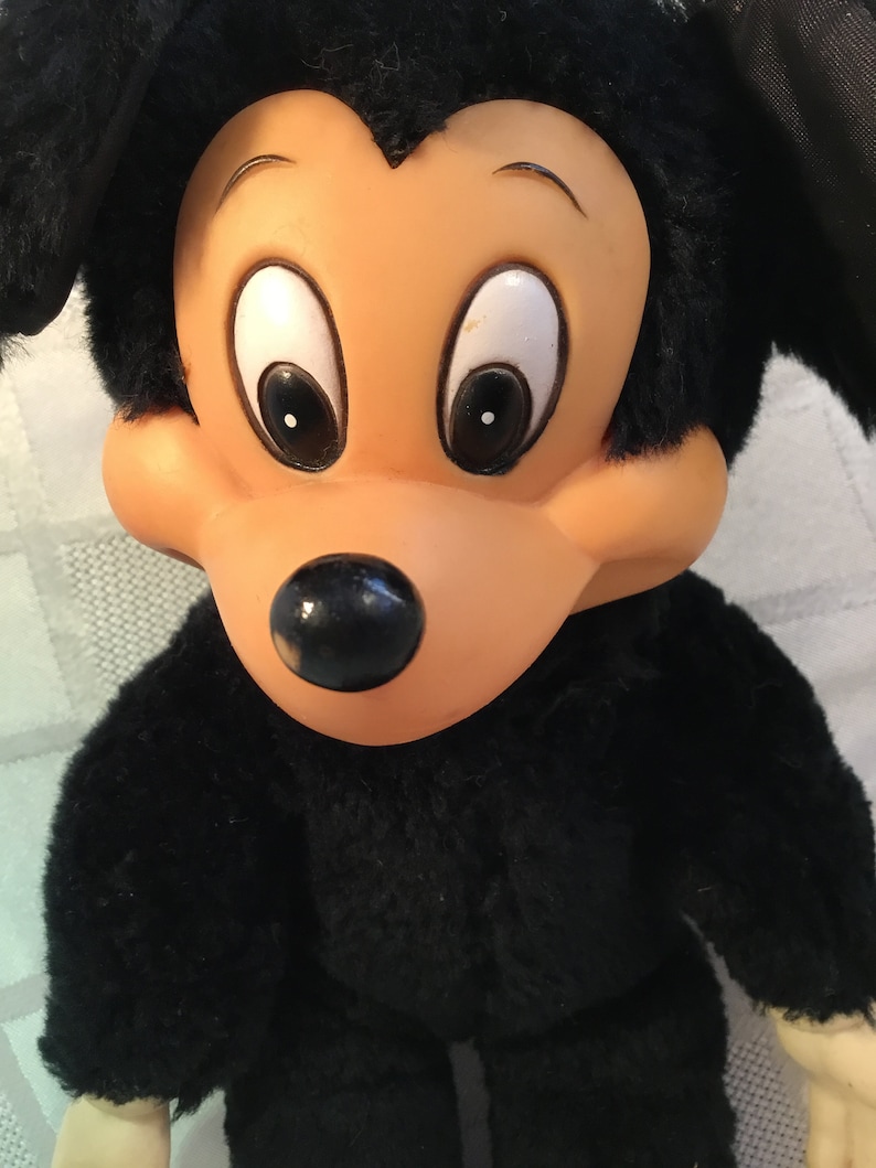 Puede incluir: Un peluche con un personaje de dibujos animados con un cuerpo negro y orejas grandes y redondas. El personaje tiene una cara bronceada con ojos grandes y blancos y una nariz negra. El pelaje del juguete es negro y suave, con una expresi&oacute;n facial detallada.