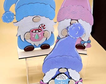 May include: Three wooden gnome figurines in blue, purple, and pink. Each gnome has a white beard, a hat, and holds a mug. One mug has a candy cane, another has a Christmas tree, and the last has a snowflake design. They stand on wooden bases.
