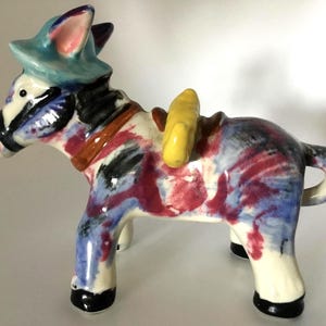 May include: A ceramic donkey figurine with a colorful, abstract design. The donkey wears a blue hat and a yellow saddle. The body is painted with red, blue, and black brushstrokes, creating a unique decorative piece.