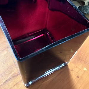 May include: A square, open-top decorative container with a glossy black exterior and a vibrant red interior. The container is made of a reflective material, and sits on a wooden surface. The container's dimensions are not visible.