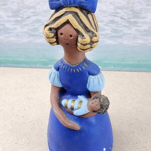 May include: A ceramic figurine of a woman in a blue dress holding a baby. The woman has brown skin, blonde hair, and a blue bow. The baby is wrapped in a blue and gold blanket. The figurine is on a sandy surface.
