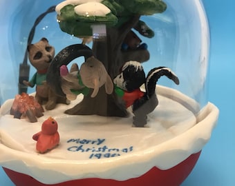 May include: A snow globe with a red base and a clear glass dome. Inside, a winter scene features a tree, various animal figurines, and the text "Merry Christmas 1996". The tree and ground are covered in snow.