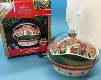 May include: Hallmark Keepsake Christmas ornament with a football stadium scene inside a clear dome. The ornament has a green base with red and gold accents. The box reads "Artists' Favorites Arctic Dome", designed by Ken Crow.