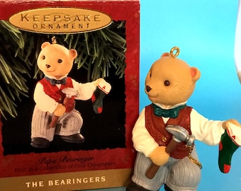 May include: A brown bear ornament wearing a red waistcoat, white shirt, and grey trousers, holding a hammer and a Christmas stocking. The figurine is part of a "Keepsake Ornament" collection. The bear has a green bow tie and black shoes.