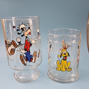 May include: Two clear glass tumblers with Disney character illustrations. One features Goofy playing golf in red, blue, and brown. The other glass has Pluto in yellow. Both glasses have a clear base.