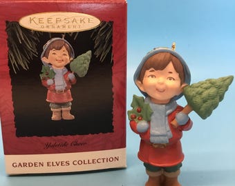 May include: A Christmas ornament from the Garden Elves Collection. The ornament shows a smiling elf in a red coat, blue scarf, and hat, holding a green Christmas tree. The box reads "Keepsake Ornament" and "Yuletide Cheer."
