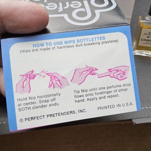 May include: A small bottle of Chanel No. 5 perfume next to a blue and white box with instructions on how to use Nips Bottlettes. The box has pink illustrations and text that reads "HOW TO USE NIPS BOTTLETTES". The perfume bottle is rectangular with a black cap.