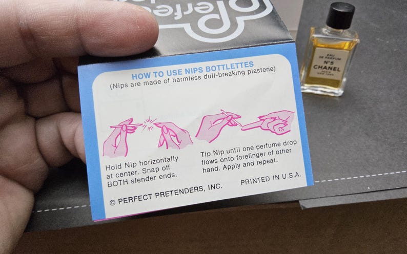 May include: A small bottle of Chanel No. 5 perfume next to a blue and white box with instructions on how to use Nips Bottlettes. The box has pink illustrations and text that reads "HOW TO USE NIPS BOTTLETTES". The perfume bottle is rectangular with a black cap.