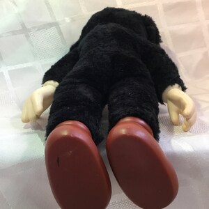 Puede incluir: Una mu&ntilde;eca de peluche vintage, vista desde atr&aacute;s, con un cuerpo negro y peludo, manos color crema y pies grandes marrones. La mu&ntilde;eca est&aacute; sobre una superficie blanca estampada. La cabeza de la mu&ntilde;eca no es visible.