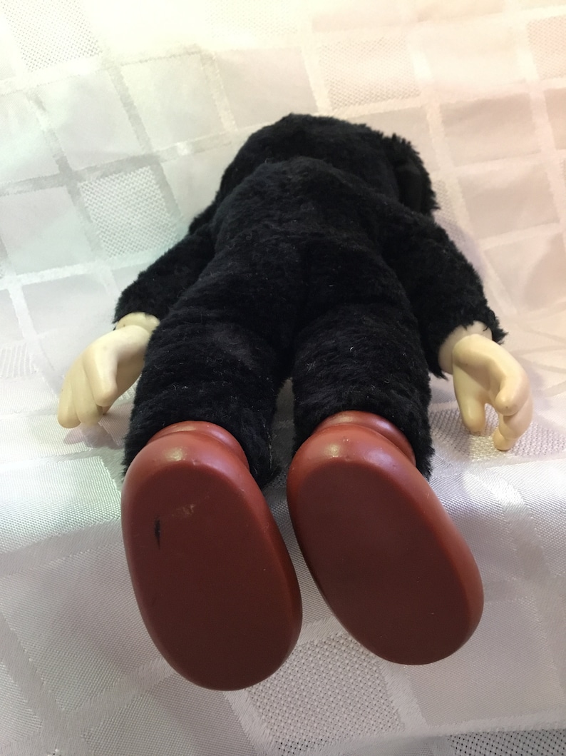 Puede incluir: Una mu&ntilde;eca de peluche vintage, vista desde atr&aacute;s, con un cuerpo negro y peludo, manos color crema y pies grandes marrones. La mu&ntilde;eca est&aacute; sobre una superficie blanca estampada. La cabeza de la mu&ntilde;eca no es visible.
