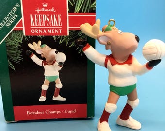May include: A Hallmark Keepsake ornament from the Collector's Series. The ornament is a reindeer wearing a volleyball uniform, holding a white volleyball. The reindeer is wearing a white shirt with red trim, green shorts, and red socks. The box reads "Reindeer Champs - Cupid."