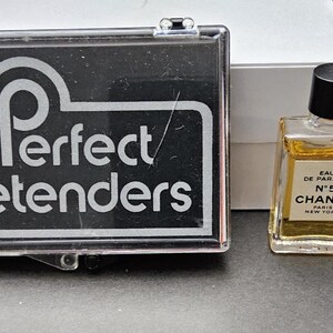 May include: A small bottle of Chanel No. 5 Eau de Parfum with a black cap, next to a black and clear case with the text "Perfect Pretenders". A white box is in the background.