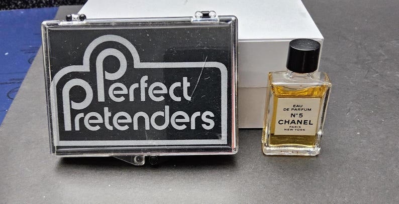 May include: A small bottle of Chanel No. 5 Eau de Parfum with a black cap, next to a black and clear case with the text "Perfect Pretenders". A white box is in the background.