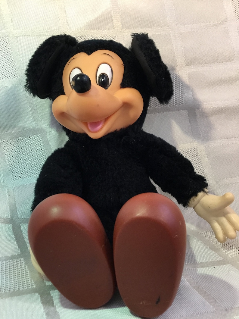 Puede incluir: Un mu&ntilde;eco de peluche vintage de Mickey Mouse. El mu&ntilde;eco tiene un cuerpo, orejas y cabeza negros, con una cara bronceada, una nariz negra y ojos blancos grandes. Tiene pies grandes, marrones y redondeados y manos color crema. El mu&ntilde;eco est&aacute; sentado sobre una superficie blanca texturizada.