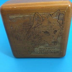 May include: Wooden box with a carved wolf design on the front. The box is rectangular with rounded edges and a light brown finish. The text "NATIONAL WILDLIFE FEDERATION" is below the wolf image.