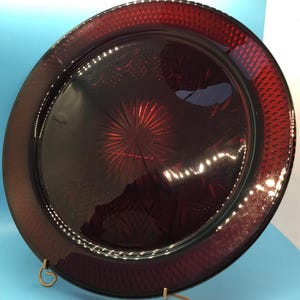 May include: A large, round, ruby-red glass plate. The plate has a textured rim and a central starburst design. The plate is likely used for serving food or as a decorative piece. The plate is displayed on a gold-coloured stand.