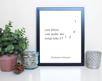 May include: A framed print with the quote "you alone can make my song take flight" from The Phantom of the Opera. The black framed print is on a white surface, with a small potted plant, a glass jar with wooden letter tiles, a pine cone, and a decorative candle holder.