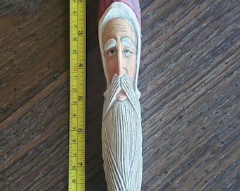 May include: A wooden Santa Claus ornament with a red hat, white beard, and a painted face. The ornament is approximately 9 inches long.
