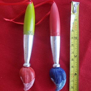 May include: Two decorative paint brush ornaments. One has a green handle and red bristles, the other has a red handle and blue bristles. Both have silver-colored metal ferrules and are approximately 4 inches long.