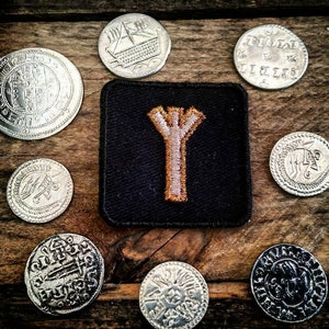 May include: An assortment of antique silver coins and a black square patch with an embroidered gold-colored rune. The coins feature various designs, including ships and intricate patterns. The items are arranged on a rustic wooden surface.