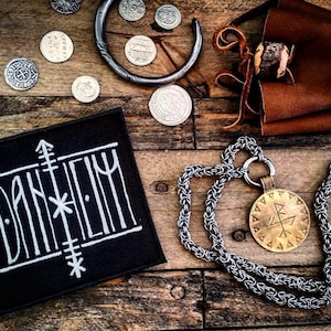 May include: Black fabric patch with white embroidery of the word "DANHELY" and a stylized symbol. The patch is on a wooden surface with a silver chain necklace and a gold pendant with a symbol.