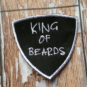 Puede incluir: Parche bordado en blanco y negro con forma de escudo con el texto "KING OF BEARDS" sobre un fondo de madera.