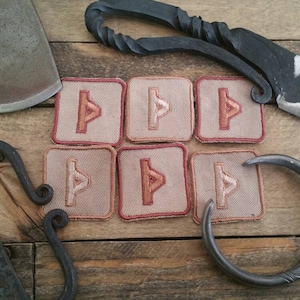 May include: Six square patches with a red and white rune symbol on a beige background. The patches are surrounded by a variety of metal tools, including a small axe, a fire starter, a knife, and a ring.