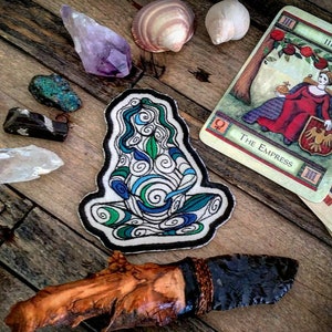 Op de afbeelding: Een blauwe en groene geborduurde patch met daarop een vrouw in een meditatieve houding met draaiende patronen. De patch is omringd door verschillende kristallen, schelpen en tarotkaarten, waaronder de kaart van de Keizerin.