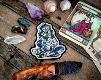 May include: A blue and green embroidered patch depicting a woman in a meditative pose with swirling patterns. The patch is surrounded by various crystals, seashells, and tarot cards, including the Empress card.