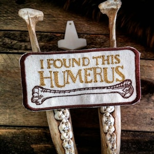 Könnte beinhalten: Ein weißer Stoff-Patch mit braunem Rand und goldener Stickerei. Der Patch lautet "Ich habe diesen Humerus gefunden" und zeigt ein stilisiertes Bild eines Knochens.