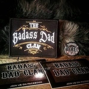 Könnte beinhalten: Ein schwarzer gestickter Aufnäher mit goldenen Nähten, auf dem "The Badass Dad Clan" steht. Es gibt auch einen kleineren Knopf mit dem gleichen Text und Design.