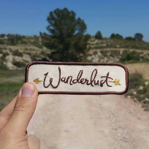 May include: A rectangular patch with the word "Wanderlust" embroidered in a dark brown script. The patch has a cream-coloured background with a brown border. Gold-coloured arrows are embroidered on each side of the word.