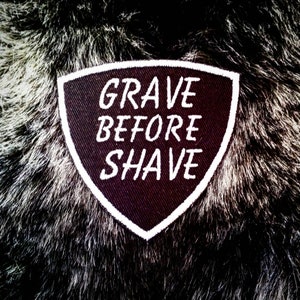 Puede incluir: Un parche de tela negro con costuras blancas en forma de escudo. El parche tiene el texto "GRAVE BEFORE SHAVE" escrito en blanco.