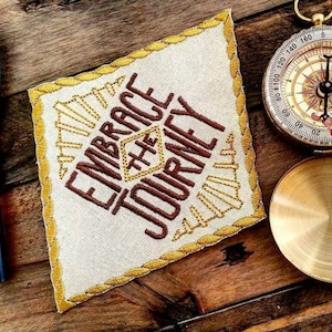 May include: A beige fabric patch with gold stitching and the words "Embrace the Journey" embroidered in brown thread. The patch is lying on a wooden surface next to a compass and a fire starter.