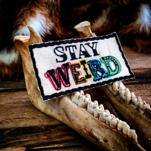 Puede incluir: Un parche blanco con el texto "STAY WEIRD" en letras negras delineadas. Las letras están rellenas de colores arcoíris. El parche está unido a un conjunto de mandíbulas de animal.