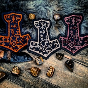 Pode incluir: Três patches bordados em forma de martelo de Thor, Mjolnir. Os patches são pretos com bordados laranja, branco e rosa. Os patches são cercados por pequenas pedras marrons de olho de tigre.