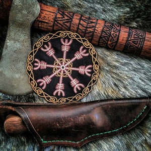 Könnte beinhalten: Eine braune Ledertasche für ein Messer mit grünen Nähten, einem braunen Holzgriff und einem schwarzen und goldenen gestickten Aufnäher mit einem rosa und weißen Symbol. Der Aufnäher ist ein kreisförmiges Design mit einem zentralen Stern und acht Speichen.