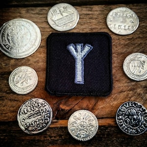 May include: A black fabric patch with a white embroidered Yggdrasil symbol, surrounded by silver coins with various designs. The coins feature Viking symbols and imagery.