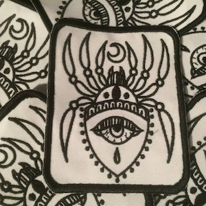 May include: Black and white embroidered iron-on patch featuring a spider with a single eye and a crescent moon above it.