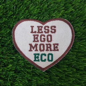 Könnte beinhalten: Ein herzförmiger Patch mit braunen Nähten und weißem Hintergrund. Der Text "Less Ego More Eco" ist in Braun und Grün gestickt.