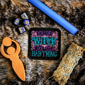 May include: A black patch with colorful embroidery that reads "You say witch like it's a bad thing". The patch is on a brown and white faux fur surface with other witchy items, including a wooden wand, blue dice with runes, and a bundle of dried sage.