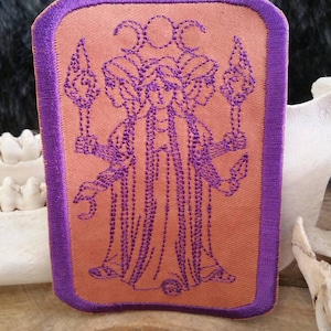 May include: A purple embroidered patch with a tan background featuring a three-headed figure with a crescent moon above each head. The figure is holding a torch in each hand.