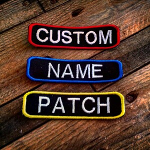 Op de afbeelding: Drie zwarte stoffen patches met witte tekst. De bovenste patch is rood en zegt "CUSTOM". De middelste patch is blauw en zegt "NAME". De onderste patch is geel en zegt "PATCH".