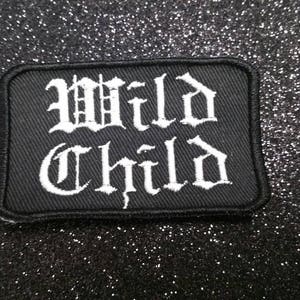 Peut inclure: Patch en tissu noir avec du texte brodé blanc qui dit "Wild Child" dans une police gothique.