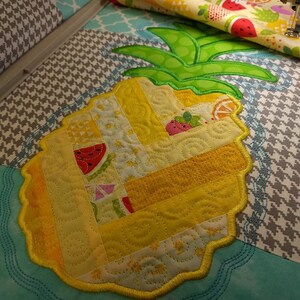 May include: A yellow pineapple-shaped quilt block with a green top and a scalloped edge. The block is made of patchwork fabric with a variety of patterns and colours, including yellow, white, and red. The block is being sewn on a sewing machine.