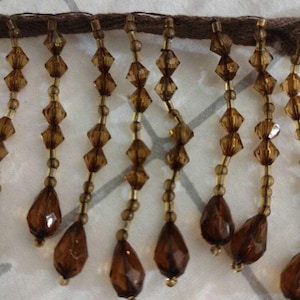 May include: Close-up of brown beaded fringe. The fringe features a dark brown woven base with strands of amber-colored beads. The beads are a mix of diamond and teardrop shapes, creating a decorative trim.
