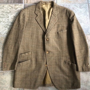 Puede incluir: Una chaqueta de tweed marrón con estampado de cuadros y cuatro botones. La chaqueta tiene dos bolsillos de solapa y un solo botón en cada puño.