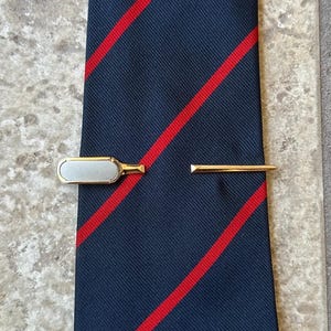 May include: A navy blue tie with red diagonal stripes, accompanied by two gold-toned tie clips. One clip features a rectangular, off-white centre, the other is a simple bar. The tie and clips are displayed on a neutral surface.